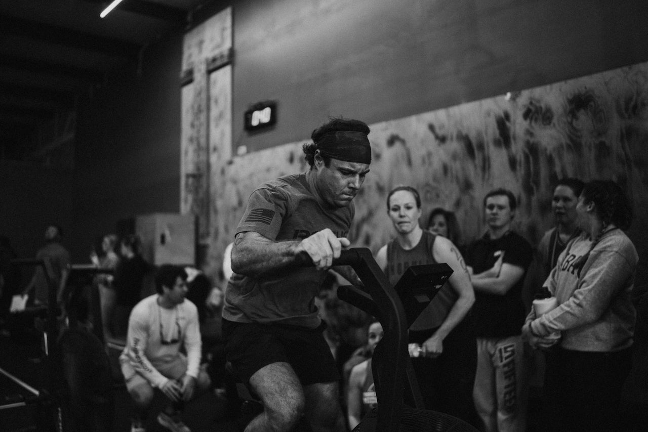 Person exercising on a crossfit machine in a gym setting with onlookers.