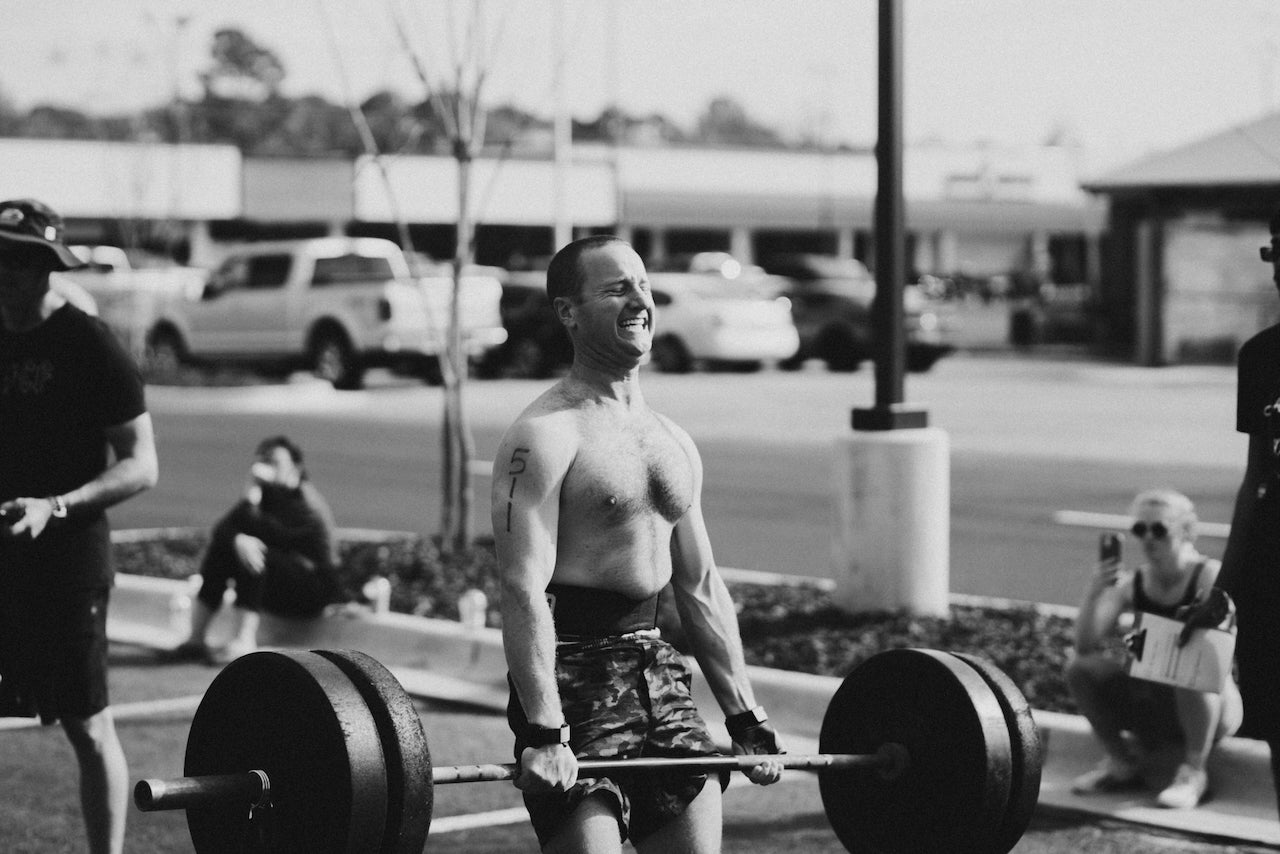 Man lifting a barbell outdoors with a parking lot and people in the background