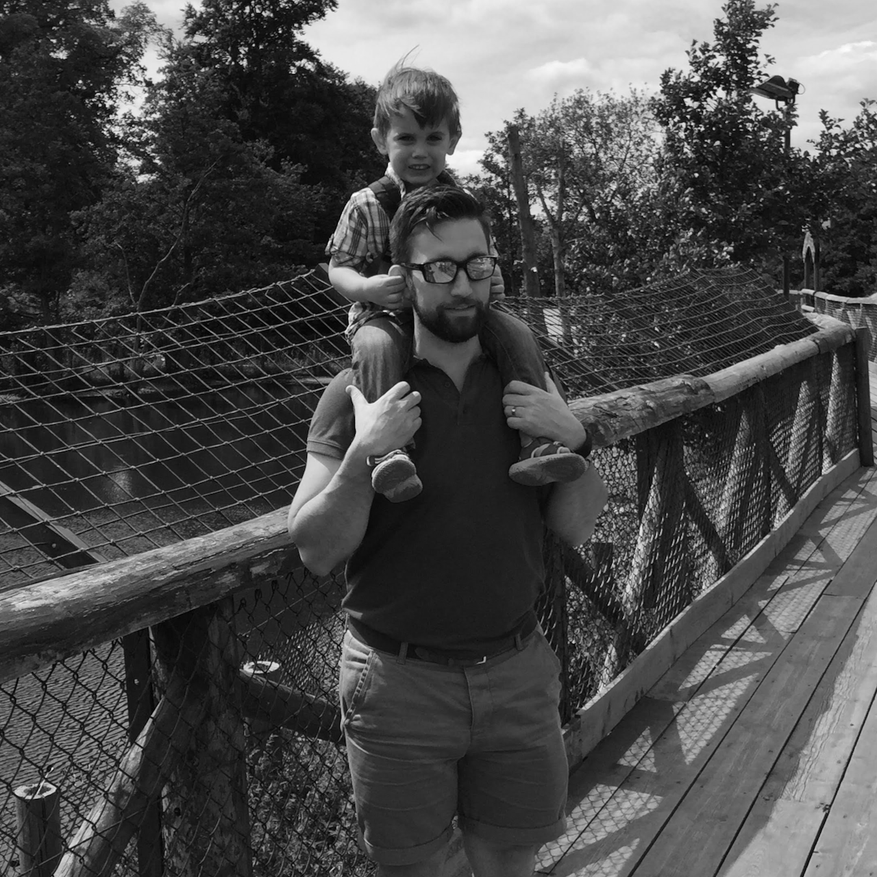 Man with a child on his shoulders standing on a wooden bridge with trees in the background